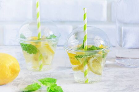 Cool Refreshing Water with Lemon and Mint Leaf. Detox and Weight Loss Concept. Healthy Organic Lemonade on White Wall Background. Slimming Drink Served in Plastic Glass with Straw Closeupの写真素材