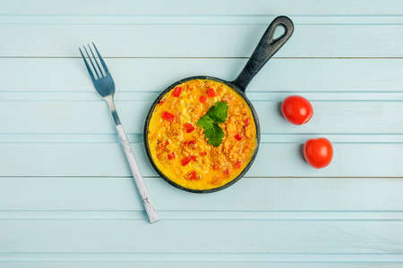 In the center of a blue background is a pan with a Turkish national recipe. Omelet made from eggs, tomato, onion and pepper, Menemen. Concept with copy space.の写真素材