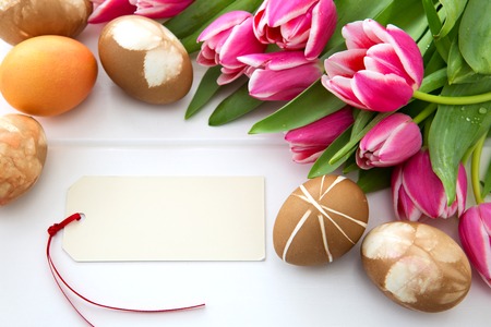 Easter card with bouquet of pink tulips and colorful eggs on white wooden .