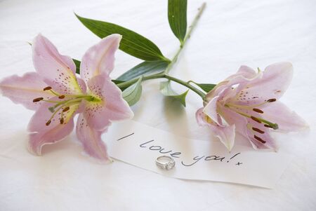 two beautiful lillies lie on the bed by a diamond engagament ring and note declaring love.の写真素材