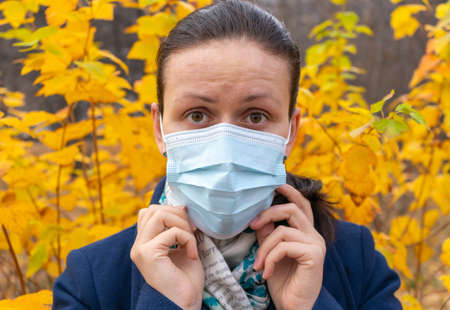 The girl adjusts the protective medical mask on her face. Autumn background in the backgroundの写真素材