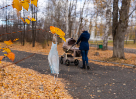 Medical face mask hangs on a branch of an autumn tree. Mom with a newborn baby in a stroller in the backgroundの写真素材