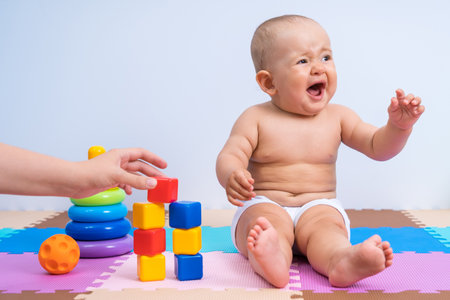 Crying child in the playroom. Mom helps a child of 8-10 months to assemble a construction set from plastic cubesの写真素材