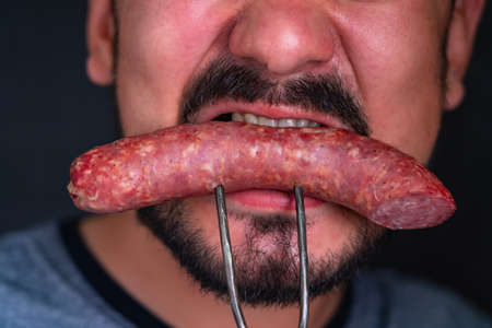 A bearded hungry man greedily eats a huge piece of sausage impaled on a fork, close-upの写真素材