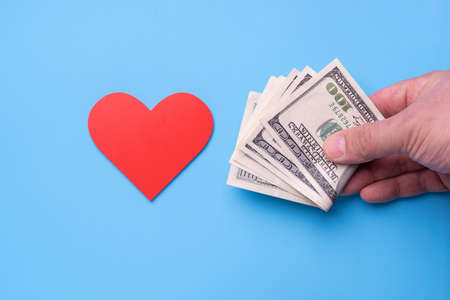 Man's hand with dollars stretches out his hand to a red heart on a blue backgroundの写真素材