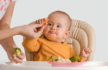 A baby aged 11-15 months takes boiled carrots from his mother's hands. Baby on a feeding chair eating boiled vegetables white backgroundの写真素材