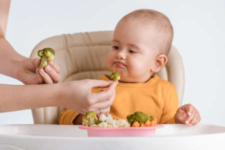 A baby aged 12-15 months curls his lips and refuses to eat boiled broccoli stretched out by his mother's handの写真素材