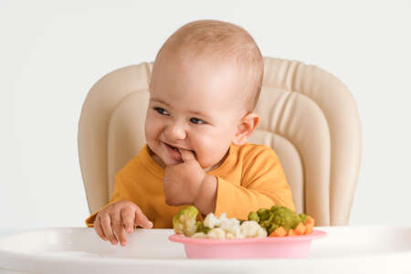 Funny baby on a feeding chair holds his fingers in his mouth. A plate of boiled vegetables in front of himの写真素材