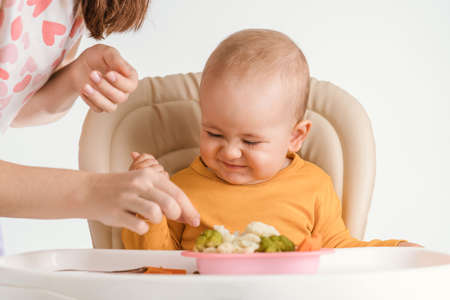 Mom feeds a one-year-old baby with boiled vegetables. A baby on a feeding chair with a funny expression on his faceの写真素材