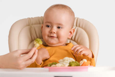 Mom's hand gives a baby sitting on a feeding chair boiled broccoliの写真素材