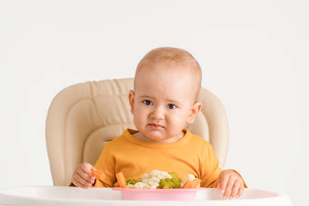 Portrait of a disgruntled baby at the dinner table. A child aged 12-17 months does not want to eat boiled vegetablesの写真素材