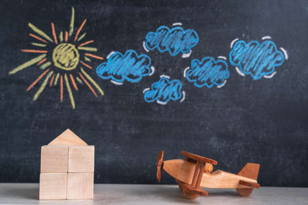 A wooden house and a toy airplane stand against a chalk board with a picture of the sun and clouds. The concept of returning home from a tripの写真素材