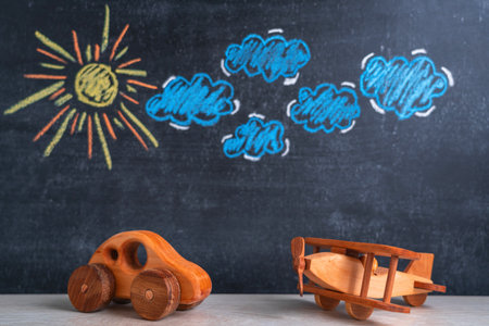 Wooden toys an airplane and a car are standing next to each other, a chalk board in the background with a picture of the sun and cloudsの写真素材