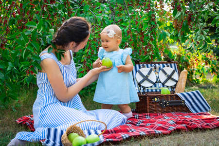 Mom with her newborn daughter at a picnic in the city park. The mother holds out a green apple to her daughter.の写真素材
