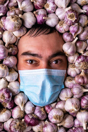 Portrait of a gloomy man in a blue medical mask among garlic bulbsの写真素材