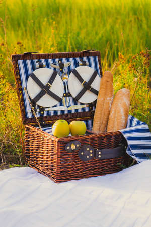 A picnic place, a wicker suitcase for picnic dishes with green apples and a baguette stands on a white blanket.の写真素材