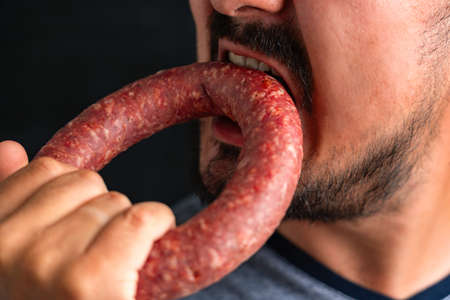 A bearded man greedily bites a round sausage. Hunger and gluttonyの写真素材
