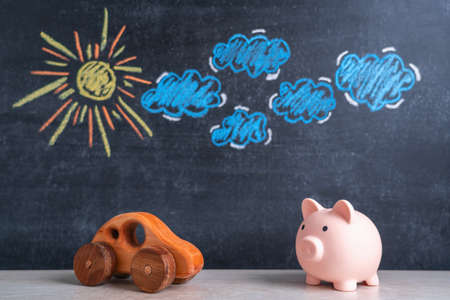 A wooden toy car and a piggy bank stand opposite each other, a chalk board in the background with a picture of the sun and clouds. The concept of car loans and the cost of car maintenanceの写真素材