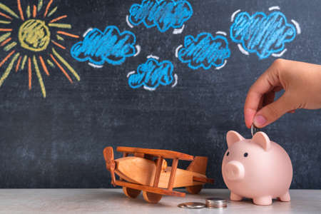 The hand puts a coin in the piggy bank. A wooden toy biplane airplane and a piggy bank stand side by side, a chalk board in the background with a picture of the sun and clouds. The concept of savings for holidays and tourist flightsの写真素材