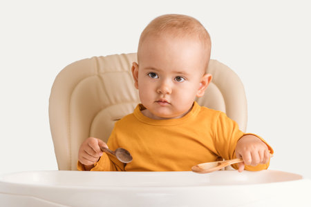A cute newborn baby aged one year is sitting on a feeding chair in orange casual clothes and holding two wooden spoons in his hands while waiting for lunch.の写真素材