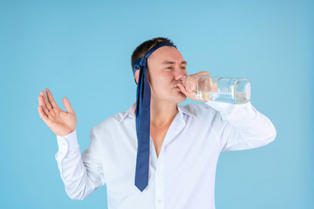 A manager with a tie on his head drinks alcohol from the neck of a bottle standing on a blue backgroundの写真素材