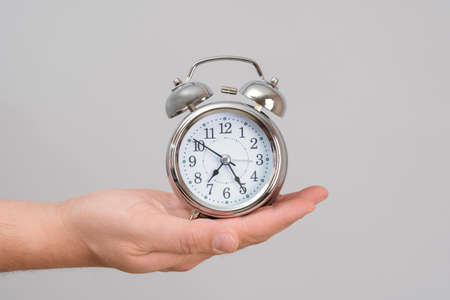 A man's hand holds a chrome alarm clock at arm's length, gray backgroundの写真素材