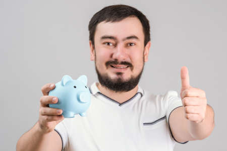 A bearded man aged 30-35 years old holds a blue piggy bank in his hand and shows a thumbs-up gesture. Close-upの写真素材