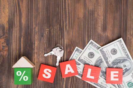The concept of selling real estate. The word SALE on red wooden cubes, keys, a small house made of wooden blocks, dollar bills on a brown background.の写真素材