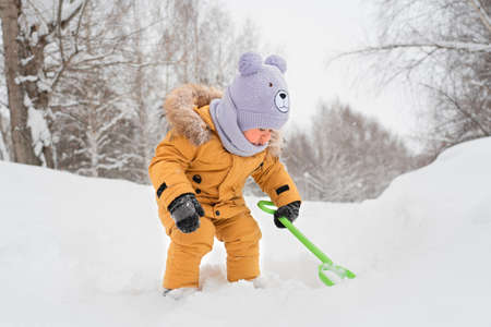 Funny toddler 12-17 months old in yellow warm clothes with a green toy shovel digging snowの写真素材