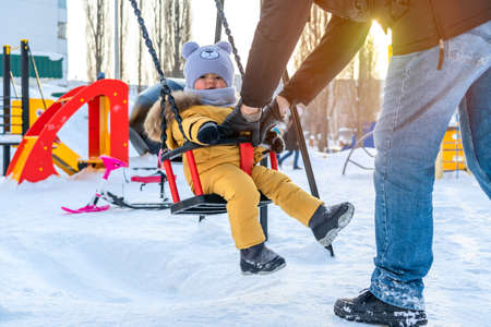 Father is rocking a toddler on a swing on a winter playground.の写真素材