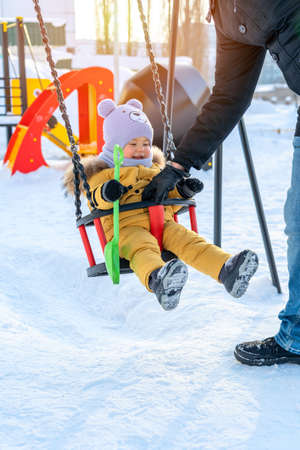 A father rocks a child on a swing on a winter playground. Active weekends in winter.の写真素材