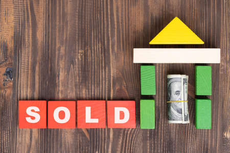 The concept of selling and buying real estate. The word SOLD on red wooden cubes. Paper dollar bills rolled up inside a wooden house made of blocks, copy spaceの写真素材