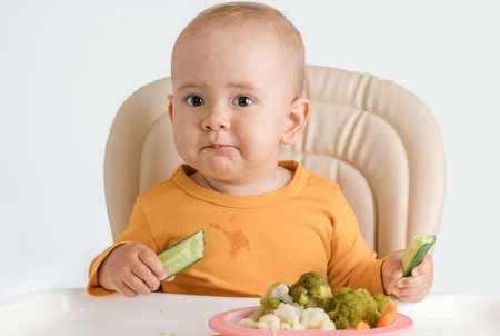 A baby on a feeding chair is eating a cucumber. The concept of proper baby food.の写真素材