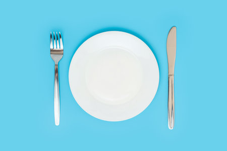 Empty white ceramic plate with a fork and a table knife on a blue background, top viewの写真素材