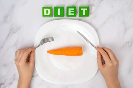 The concept of a vegetable diet. Female hands with a fork and a table knife over an orange carrot lying on a white plateの写真素材