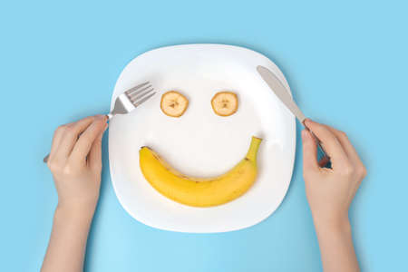 The concept of a positive attitude to diet and healthy eating. Women's hands with cutlery over a dish with a banana on a blue background, top viewの写真素材