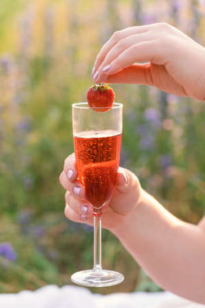 A woman's hand puts a strawberry berry in a glass of sparkling wine at a picnicの写真素材