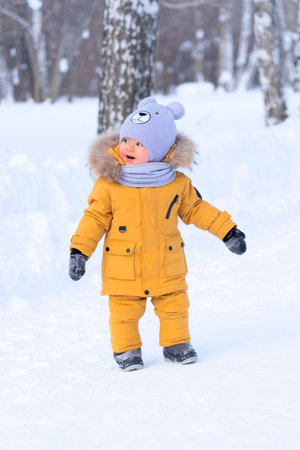 Portrait of a toddler 15-20 months old in yellow warm clothes looking back standing on a snowy path in the parkの写真素材