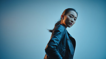 A young Asian woman dressed in a dark leather jacket, against a blue studio background, with confident expression.の素材