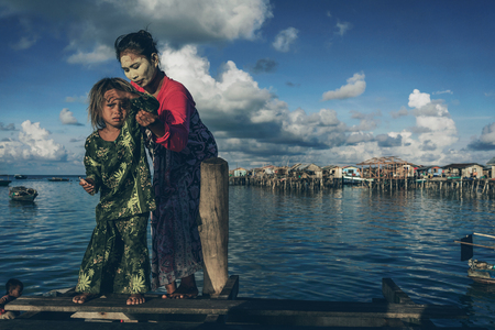 Denawan Island, Sabah Malaysia - April 30, 2016 : A mother helping her child to stand on a broken wooden bridge. Both were wearing a traditional clothing with the mother had a natural paste on her face as the natural sun block.のeditorial素材
