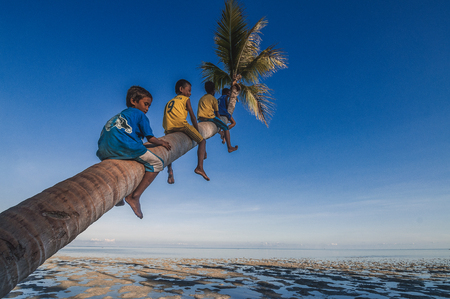 Denawan Island, Sabah Malaysia - April 29, 2016 : A goup of local kids siting on coconut tree's trunk overlooking the low tide ocean over a clear blue sky.のeditorial素材