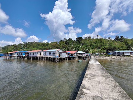 Sabah, Malaysia - January 27th, 2026: A wide-angle view of a traditional water village set against a lush, green hillside. A series of wooden houses elevated on stilts line the coast, connected by a long, weathered concrete pier that leads the eye into thのeditorial素材