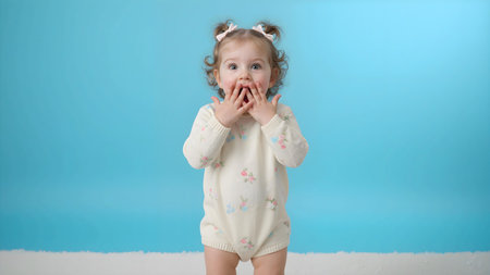 Cute toddler girl child with surprised facial expression, standing against bright blue backgroundの素材