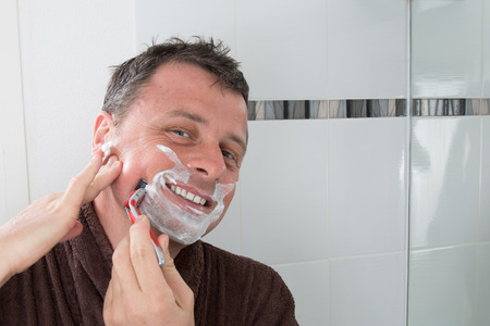 Man shaving with a razor blade and shaving cream in bathroomの写真素材