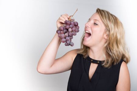 A beautiful young blond girl eating a bunch of black grapesの写真素材