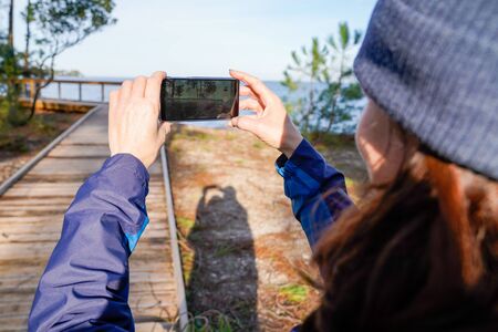 back view tourist woman take phone photo picture in pine forest wooden pontoon lake of Carcans Franceの写真素材
