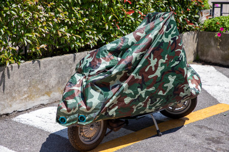 motorcycle scooter protected by camouflage military tarpaulin protective under cover in city street motorbike with jacketの写真素材