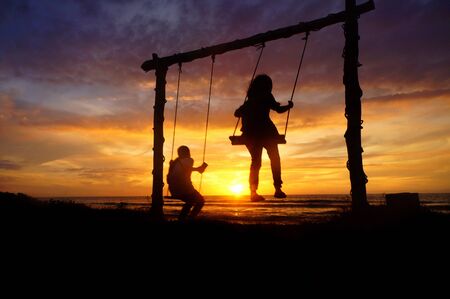 Slightly blur and grainny silhouette image of two girls on a makeshift swing by the beach during a...
