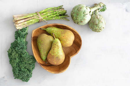 Heart-shaped coconut wood bowl with pears, kale leaves, asparagus and artichokes. Still life with winter fruit and vegetables. Green energy. Healthy dietの写真素材