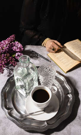 Mysterious woman drinking black coffee with glass and jug of water on elegant silver tray with dark...